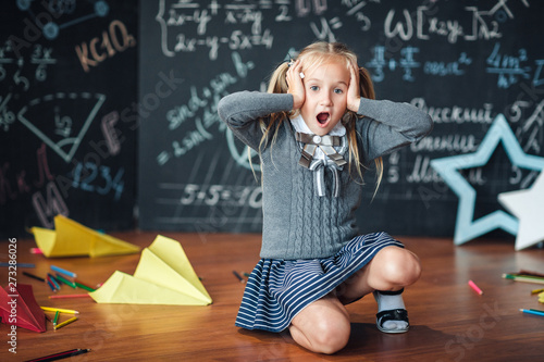 Little girl blonde in school uniform holding hands on her head., open his mouth against chalkboard with school formulas. Complex school program, does not match the age of the child.