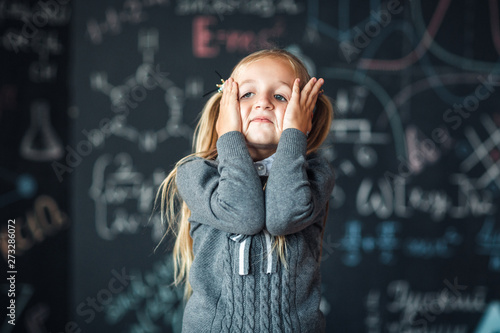 Portrait of little blond girl, grabbing her head. chalkboard with school formulas at the background, concept photo
