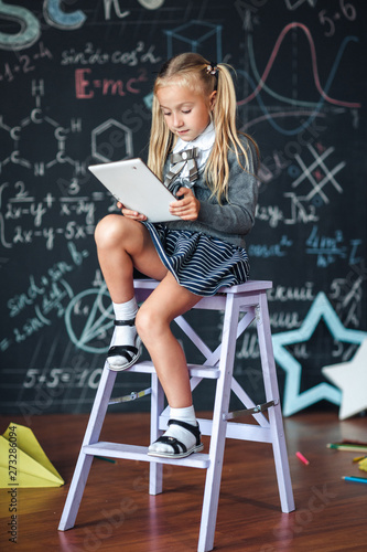 Little blond Girl in school uniform holding white tablet pc in chemistry class. Chalkboard with school formulas background.