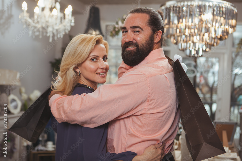 Fototapeta premium Happy mature couple smiling to the camera, holding shopping bags. Handsome bearded mature man embracing his beautiful wife while shopping together. Retail, consumerism, sales concept