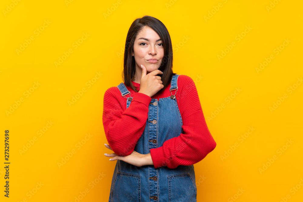 Young Mexican woman with overalls over yellow wall thinking Young Mexican woman with overalls over yellow wall thinking
