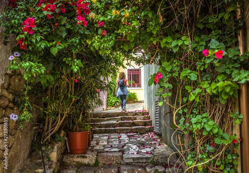 Beautiful narrow street with flowers in Plaka district, Athens, Greece. Scenic alley like overgrown tunnel in the Athens center. Young woman in the distance. Concept of travel and vacation in Athens.