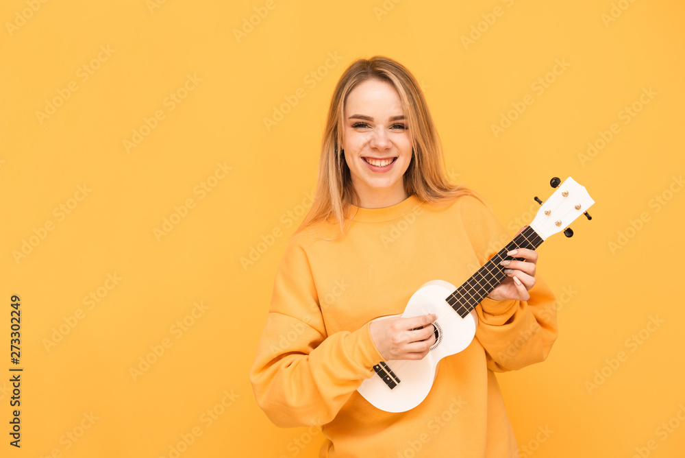 Portrait of a smiling girl in bright clothes, standing with a Hawaiian guitar in her hands on a yellow background,looking in camera and rejoicing. Blonde musician plays ukulele and laughs, isolated. Portrait of a smiling girl in bright clothes, standing with a Hawaiian guitar in her hands on a yellow background,looking in camera and rejoicing. Blonde musician plays ukulele and laughs, isolated.