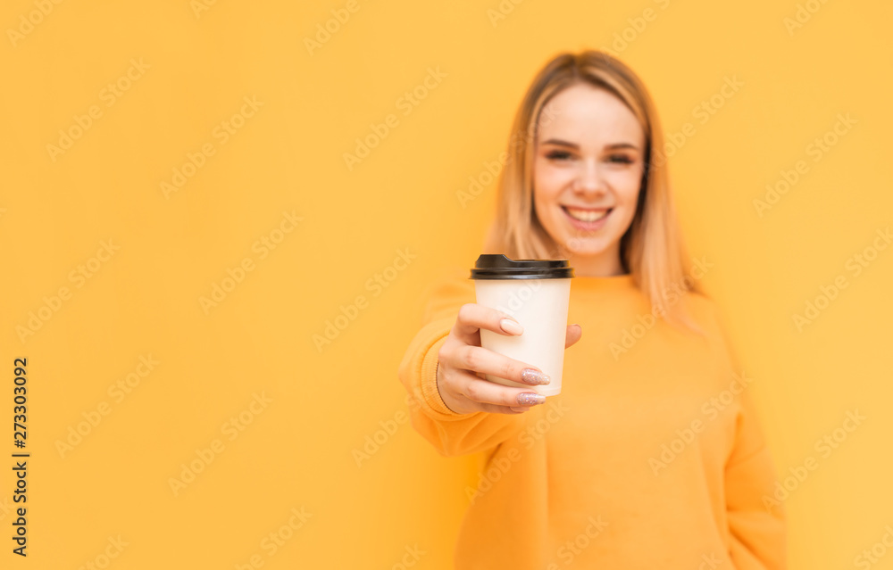 Φωτογραφία Smiling blonde in an orange sweater shows a cup of coffee in the camera on a yellow background, looking into the camera