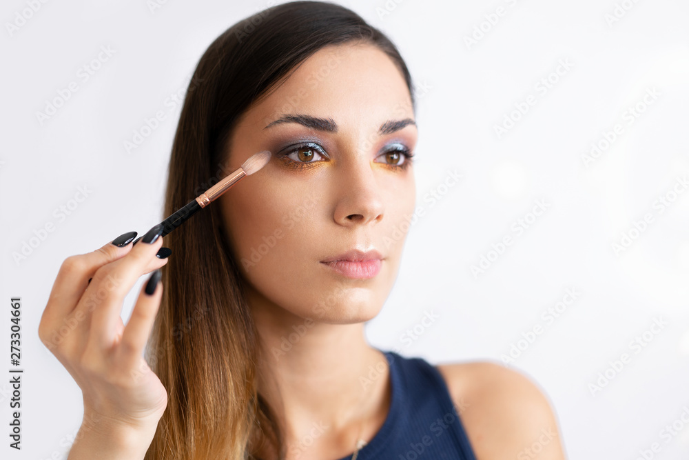 Make-up artist applying bright base color eyeshadow on model's eye and holding a shell with eyeshadow on background