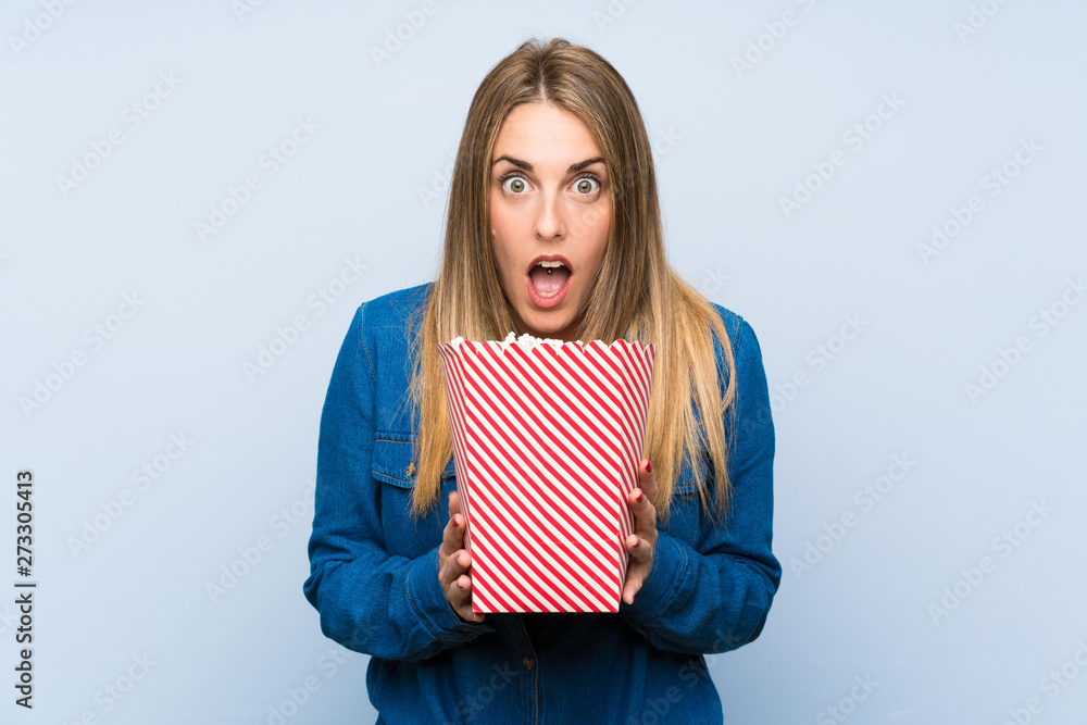 Blonde woman with popcorns over blue wall doing surprise gesture Blonde woman with popcorns over blue wall doing surprise gesture