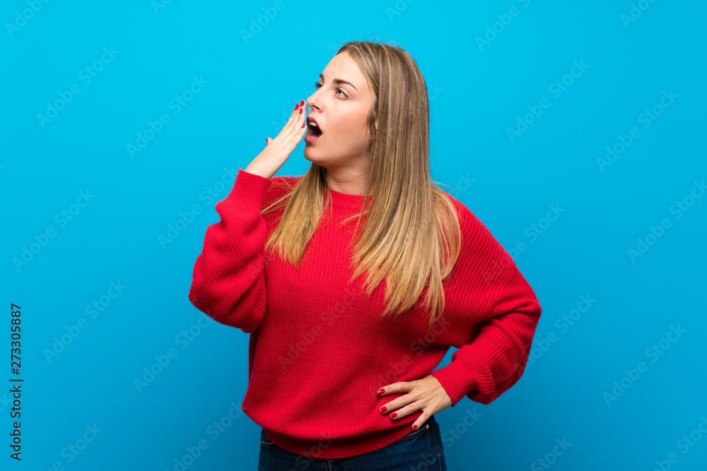 Woman with red sweater over blue wall yawning and covering wide open mouth with hand Woman with red sweater over blue wall yawning and covering wide open mouth with hand