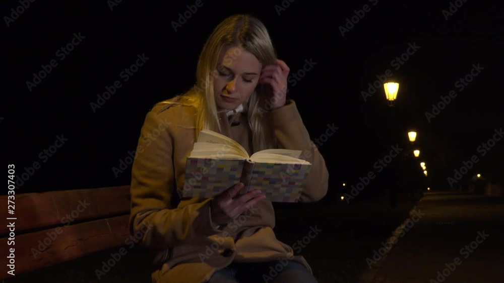 A young beautiful woman sits on a bench and reads a book in an urban area at night - a line of streetlights in the background A young beautiful woman sits on a bench and reads a book in an urban area at night - a line of streetlights in the background