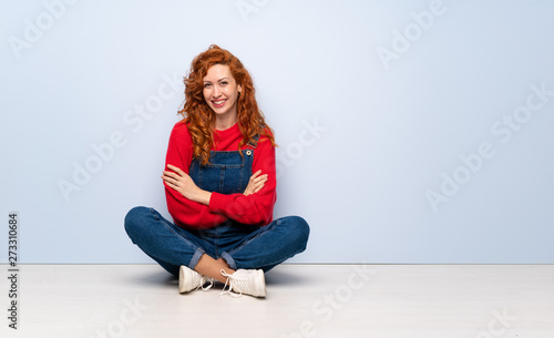 Redhead woman with overalls sitting on the floor looking up while smiling