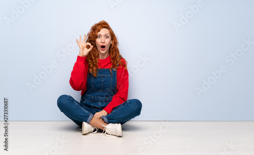 Redhead woman with overalls sitting on the floor surprised and showing ok sign