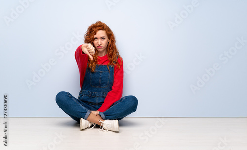 Redhead woman with overalls sitting on the floor showing thumb down with negative expression