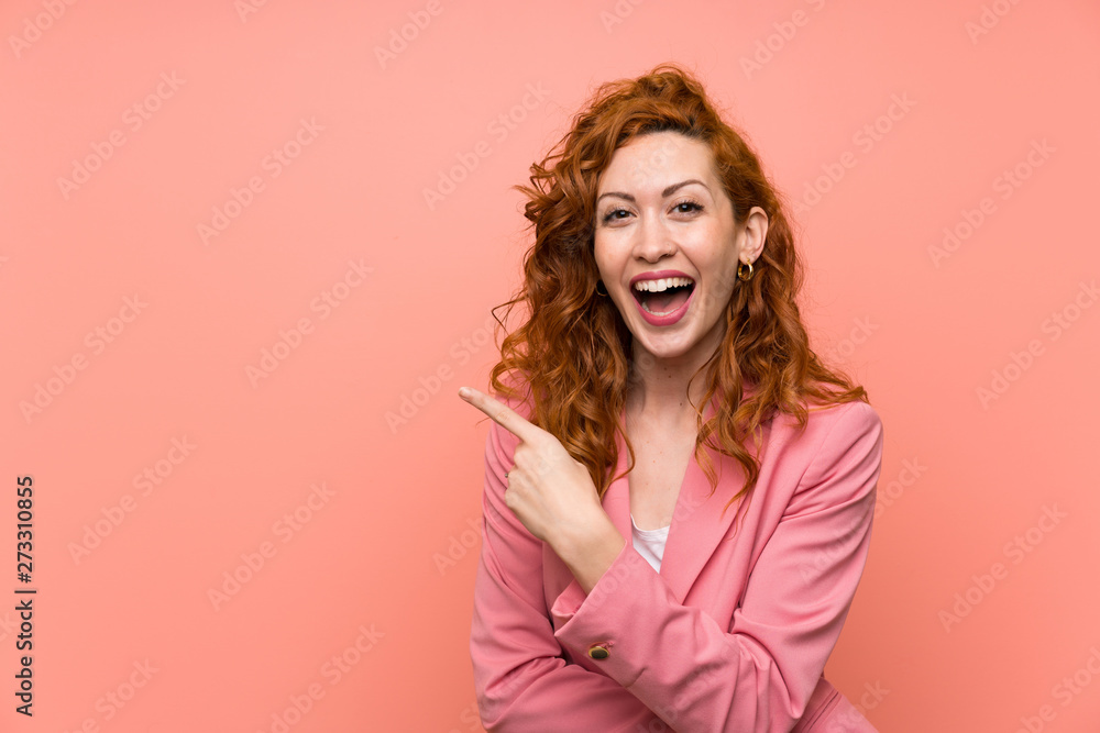 Redhead woman in suit over isolated pink wall pointing finger to the side Redhead woman in suit over isolated pink wall pointing finger to the side
