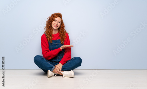 Redhead woman with overalls sitting on the floor presenting an idea while looking smiling towards