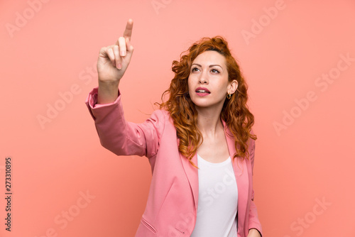 Redhead woman in suit over isolated pink wall touching on transparent screen photo