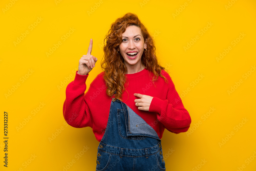 Redhead woman with overalls over isolated yellow wall with surprise facial expression Redhead woman with overalls over isolated yellow wall with surprise facial expression