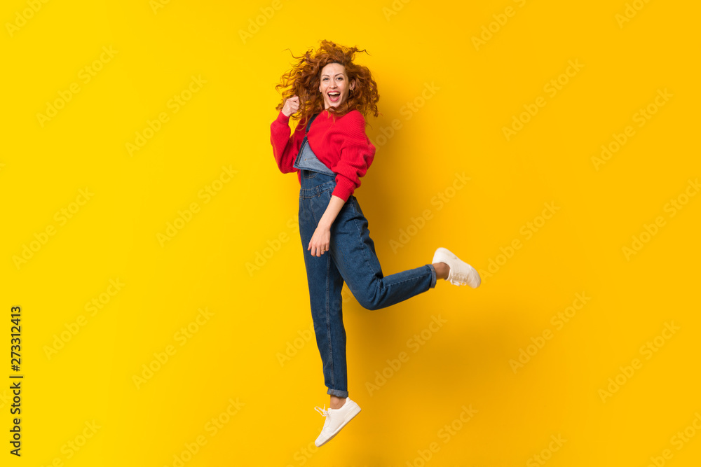 Redhead woman with overalls jumping over isolated yellow wall