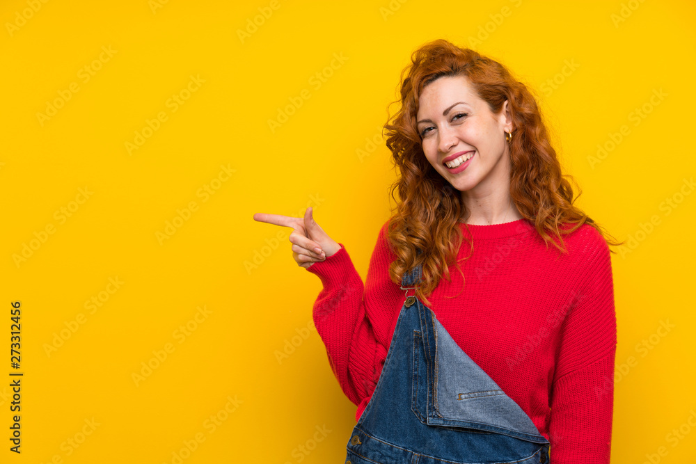 Redhead woman with overalls over isolated yellow wall pointing finger to the side Redhead woman with overalls over isolated yellow wall pointing finger to the side