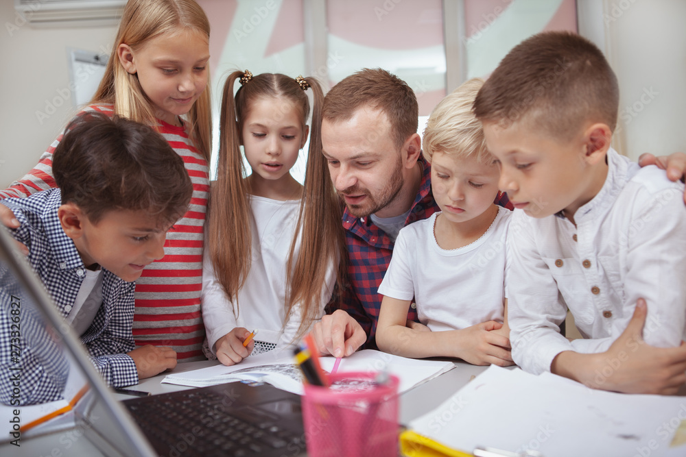 Mature male teacher enjoying working with kids at primary school, helping them with a project. Friendly teacher explaining tasks to his little students at elementary school