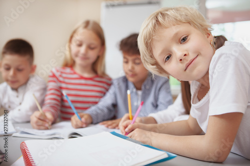 Close up of a cute little girl smiling to the camera while drawing at art class with her friends on background, copy space