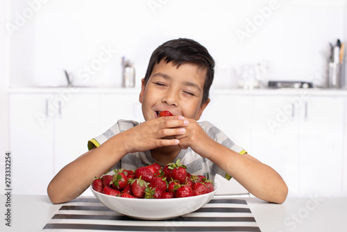 Shot of a smiling boy sitting in the kitchen eating strawberries with closed  eyes from pleasure.