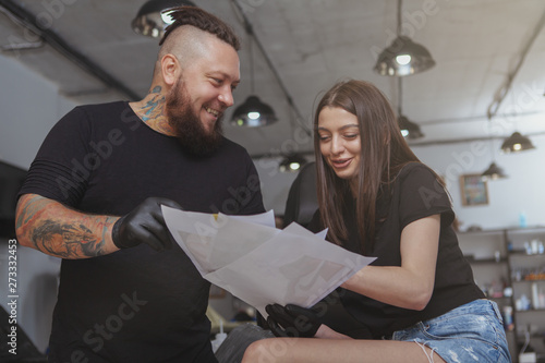 Cheerful bearded tattoo artist laughing, talking to his female client while showing her tattoo sketches. Beautiful woman choosing new tattoo sketch at tattoo salon