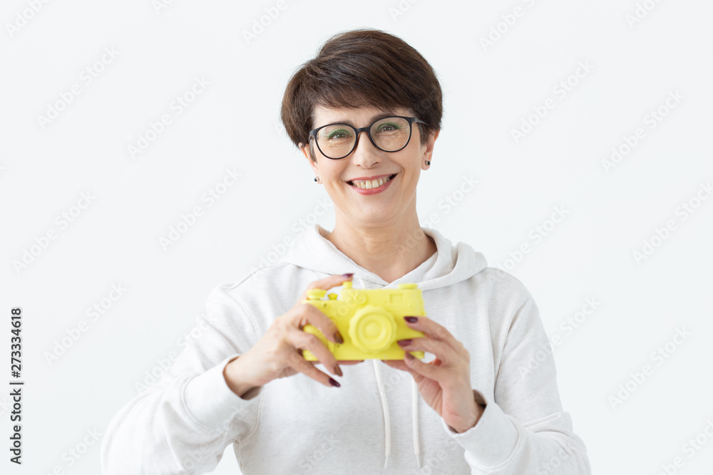 Positive middle-aged woman in glasses holds in hand a vintage yellow camera posing on a white background. Concept of photo lovers and hobbies Positive middle-aged woman in glasses holds in hand a vintage yellow camera posing on a white background. Concept of photo lovers and hobbies