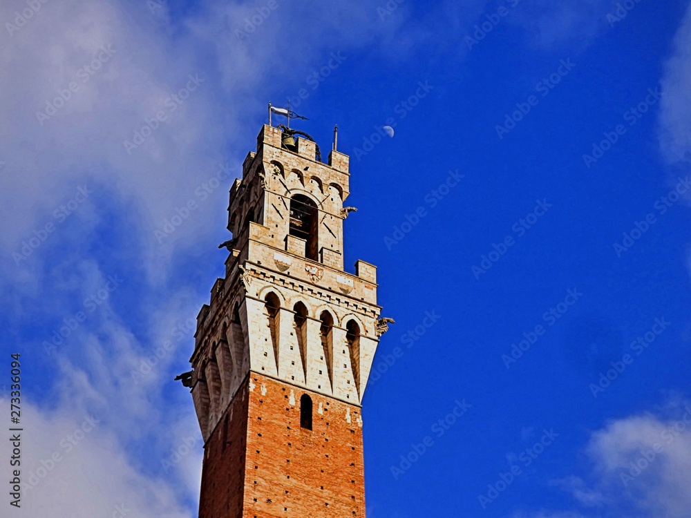 Looking up at the bell tower of a medical cathedral in a Tuscan hill town of Italy. Blue sky and white cloud background with the moon showing in the daytime.