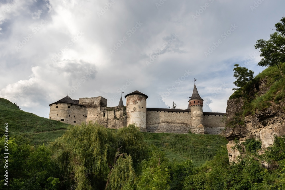 Part of the medieval Kamianets-Podilskyi fortress of the XVI century against the background of greenery and rocks in the foreground in the cloudy sky. Ukraine.