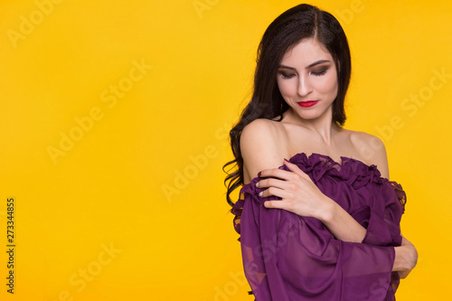 Beautiful brunette woman in a purple dress and bare shoulders on a yellow background. Copy space.