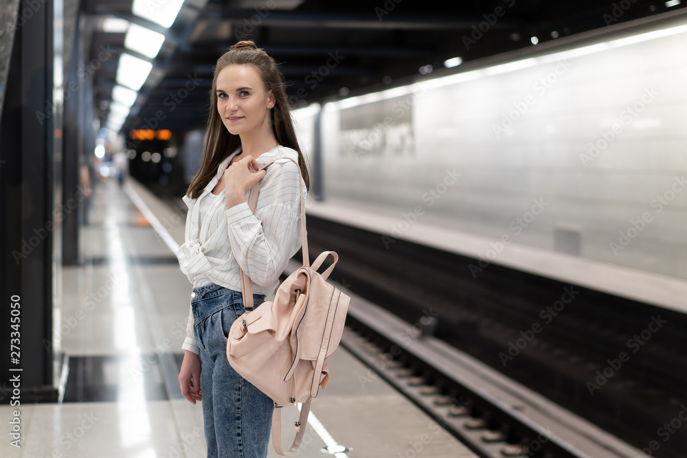 Fototapeta premium European girl with a backpack standing at the subway station and waiting for the train. The train is out of focus to convey the effect of the reality of the movement. The main object is a girl.