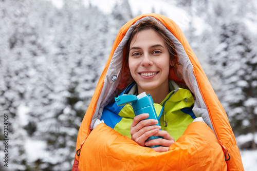 Horizontal view of smiling attractive tourist feels cold after expedition in snowy mountains, wrapped in sleeping bag, drinks hot tea, poses over mountanious forest covered with snow. Enjoying nature photo