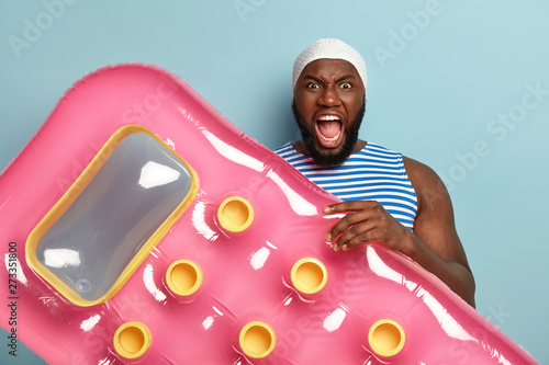 Irritated young Afro man holds air mattress bed, yells with annoynace, being angry with other vacationists on beach, has spoiled pool party, cannot swim in good place, expresses negative emotions photo