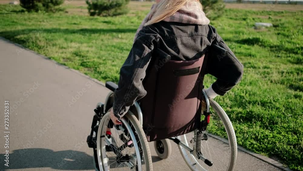 Woman is using wheelchair for moving outdoors in sunny day, back view Woman is using wheelchair for moving outdoors in sunny day, back view