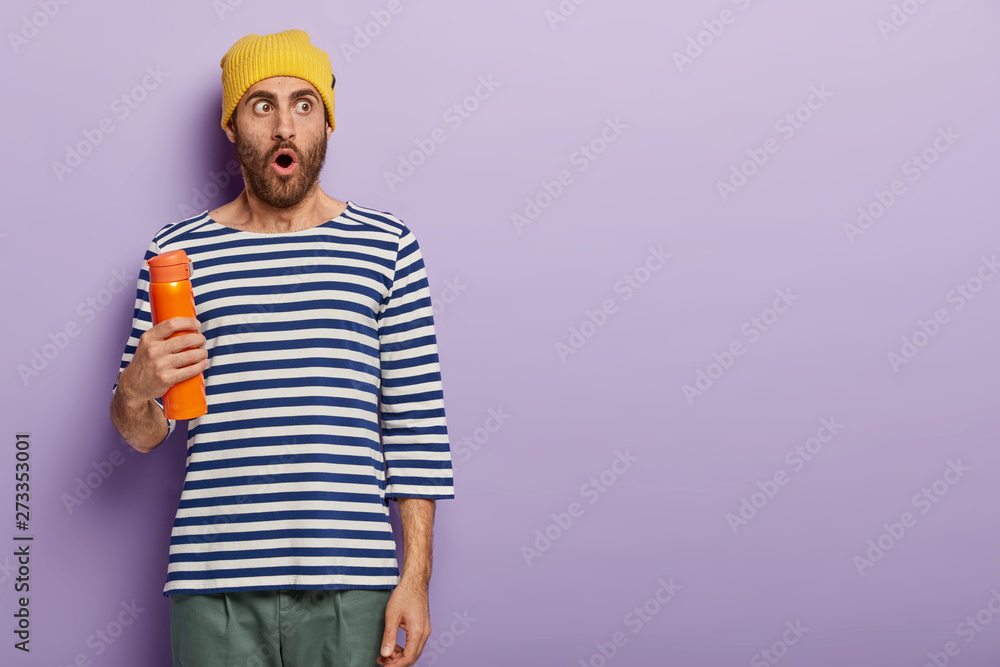 Studio shot of impressed student drinks hot coffee from thermos bottle during break, focused aside with astonishment, dressed in stylish apparel, models against purple wall, copy space for text