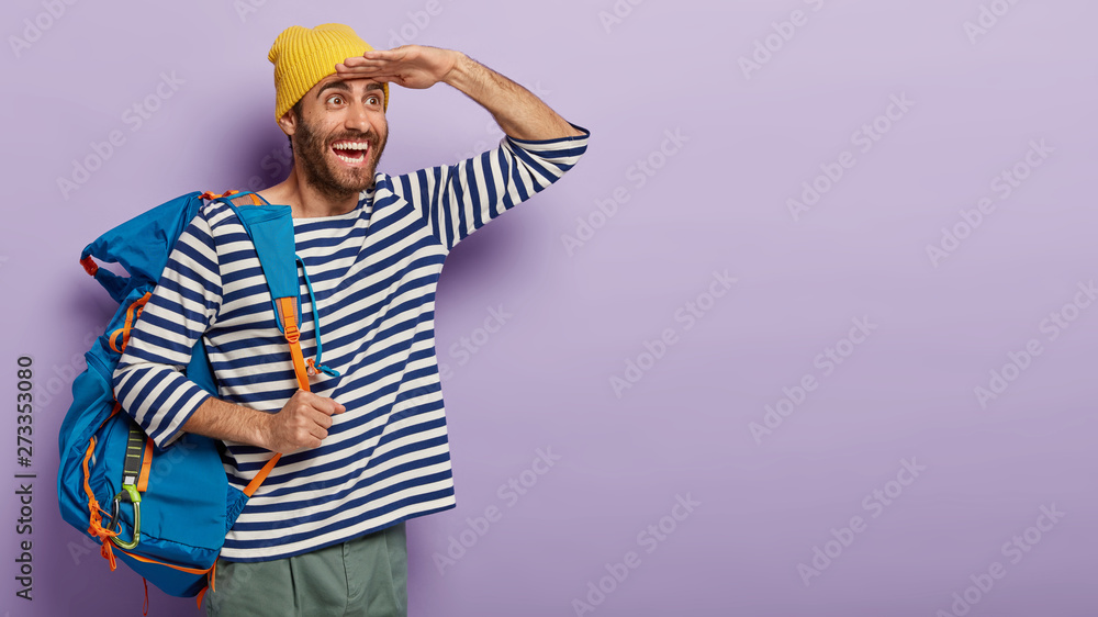 Cheerful traveler keeps palm near forehead, focuses gladfully into distance, notices place he aimed to reach, carries big tourist backpack, wears stylish yellow hat and striped jumper. People, hiking Cheerful traveler keeps palm near forehead, focuses gladfully into distance, notices place he aimed to reach, carries big tourist backpack, wears stylish yellow hat and striped jumper. People, hiking