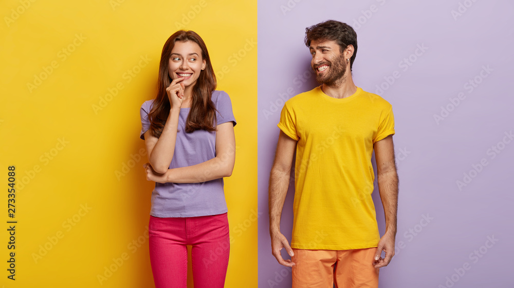 Optimistic young female and male with broad smiles, feel happy, dressed in casual clothes, have fun together, stand against purple and yellow background. People, relations, togetherness concept - obrazy, fototapety, plakaty 
