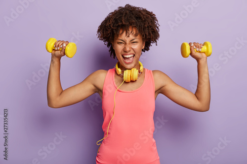 Horizontal shot of energized happy Afro woman raises arms with dumbbells, enjoys sport training with music in headphones, dressed in casual pink vest, poses indoor. People, mood, sport, motivation