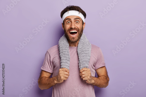 Happy male runner smiles broadly, surprised to cover long distance on marathon, has towel on neck, wears casual t shirt and white headband, isolated over purple background. Sport and training concept