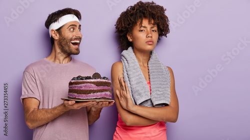 Beautiful confident sport girl with Afro haircut refuses eating sweet cake, shows no gesture, supports healthy nutrition and lifestyle. Happy man wears headband and t shirt suggests dessert to woman