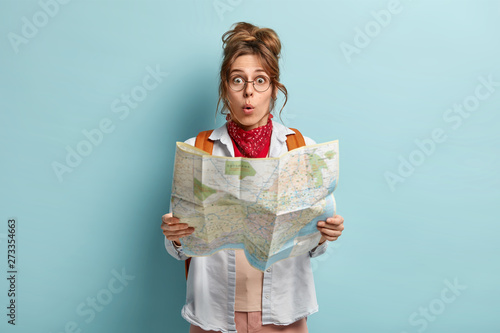 Shocked female European tourist has tour around world, surprised to loose way, reads map, wears round spectacles, carries backapck, poses over blue background. Summer travel concept. Sightseeing photo