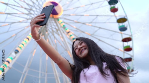 Asian beautiful woman using smartphone doing selfie at amusement park carnival near ferris wheels. Woman happy travel night funfair festival 4k resolution.