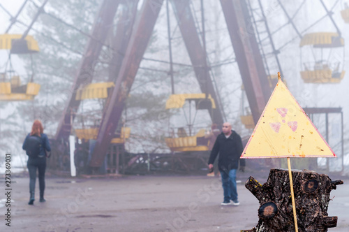 Ukraine, Pripyat, Chernobyl zone of alienation - 31.03.2018: soft focus of tourists in fog ruined park near ferris wheel and yellow warning of radiation danger sign 