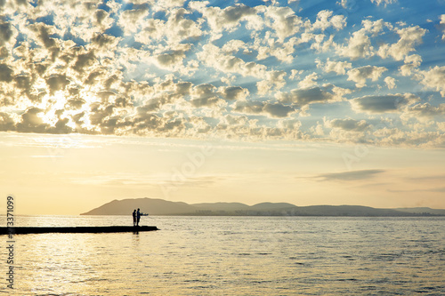 Silhouette of two people on the pier fishing in the sea at sunset against the mountains