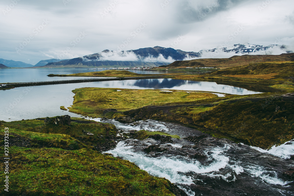 Spectacular view of amazing Icelandic landscape from Kirkjufellsfoss on Snaefellsnes peninsula. Clear water of the stream, green grass and moss, lake, blue mointains, and gray clouds.