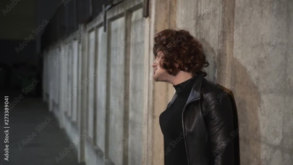 Young caucasian funny guy actor dressed like woman, wearing black leather jacket with blouser and put on head brown wig, sings outside standing next to beige wall in street in the evening. Young caucasian funny guy actor dressed like woman, wearing black leather jacket with blouser and put on head brown wig, sings outside standing next to beige wall in street in the evening.