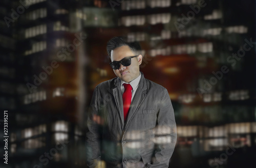 A young business man in corporate attire with red tie and sunglasses simply standing behind a window glass with the city light reflections. A man looking out the window at night.