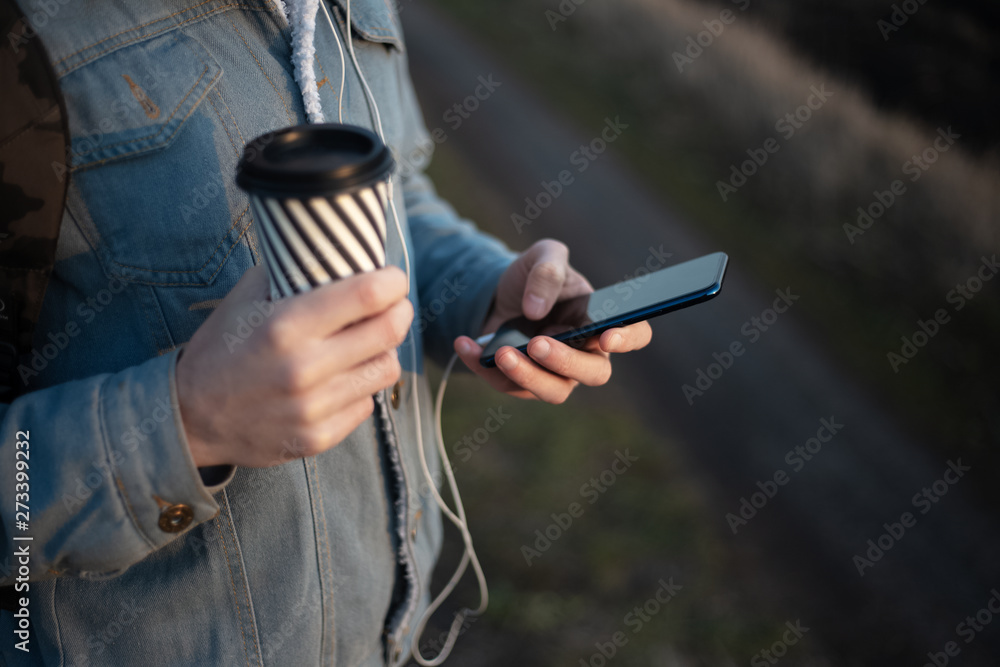 Close-up of hands of young modern guy with backpack, holding the coffee cup and using smartphone with earphones