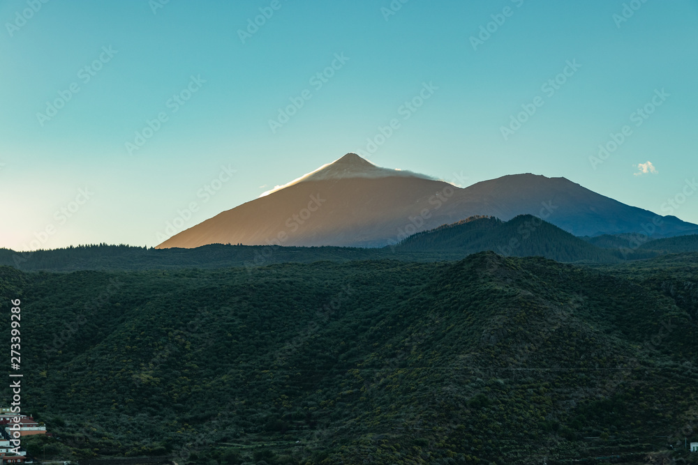 Mountain Teide at early morning. Top of volcano partly covered by the creeping thin clouds. Sunrise. Bright blue sky. Green hills on the foreground. Tenerife, Canary Islands, Spain