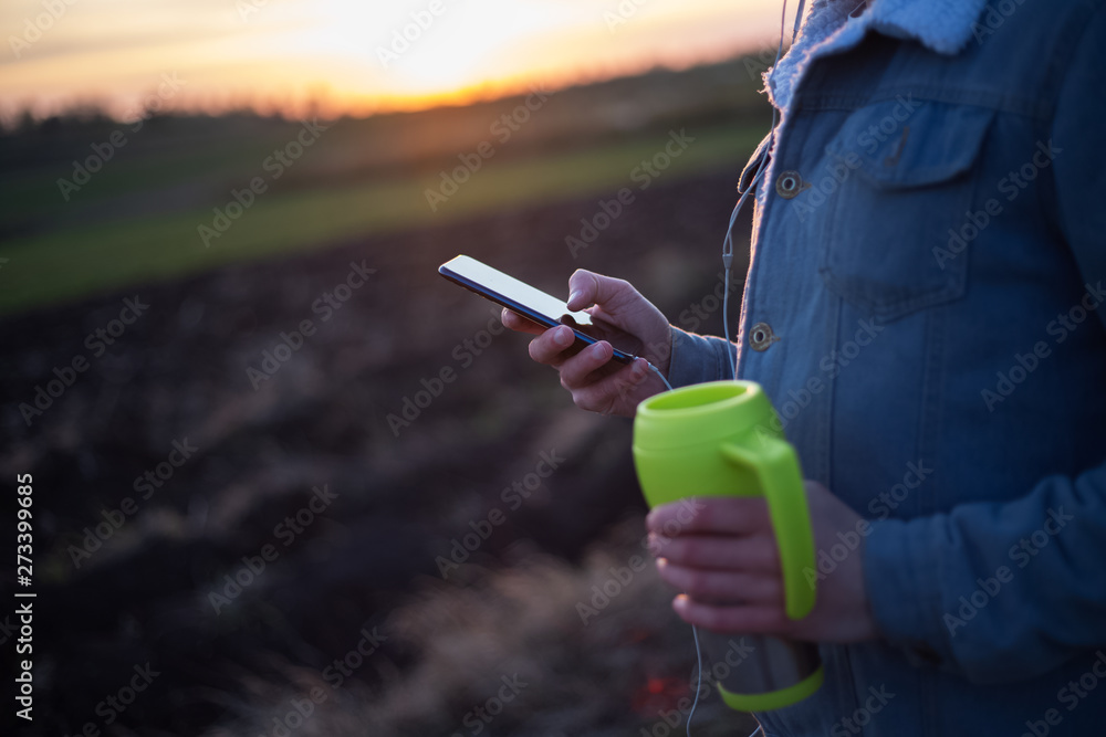 Close-up of green mug with coffee and smartphone in hands of man at outdoor sunset