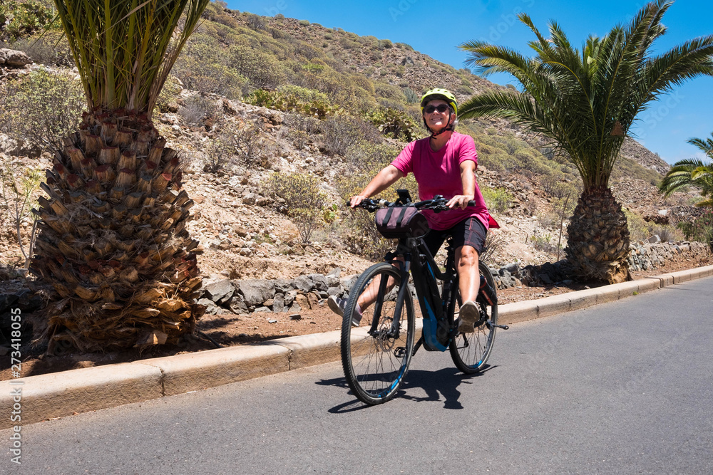 Fototapeta premium Smiles and happiness of a senior lady bicycling with an e bike. Blue sky and palm trees on background. Tropical climate